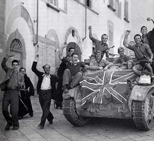 immagine storica in bianco e nero legata all'esultanza collettiva per la liberazione della città di Arezzo durante la Seconda Guerra Mondiale. Al centro e sulla destra domina un mezzo corazzato cingolato, sul cui fronte è chiaramente visibile e stesa una grande bandiera del Regno Unito. Sopra e a bordo del mezzo militare ci sono diversi soldati in divisa.